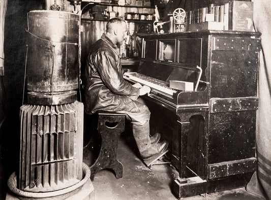 Classic Herbert Ponting photograph from Captain Scott's Terra Nova expedition (1910-1913), showing Cecil Meares sitting at the pianola in The Hut.