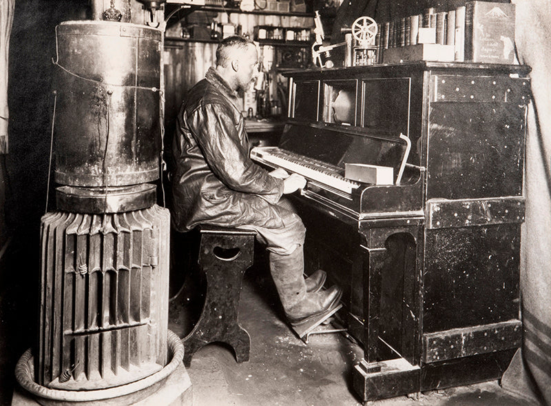 Classic Herbert Ponting photograph from Captain Scott's Terra Nova expedition (1910-1913), showing Cecil Meares sitting at the pianola in The Hut.